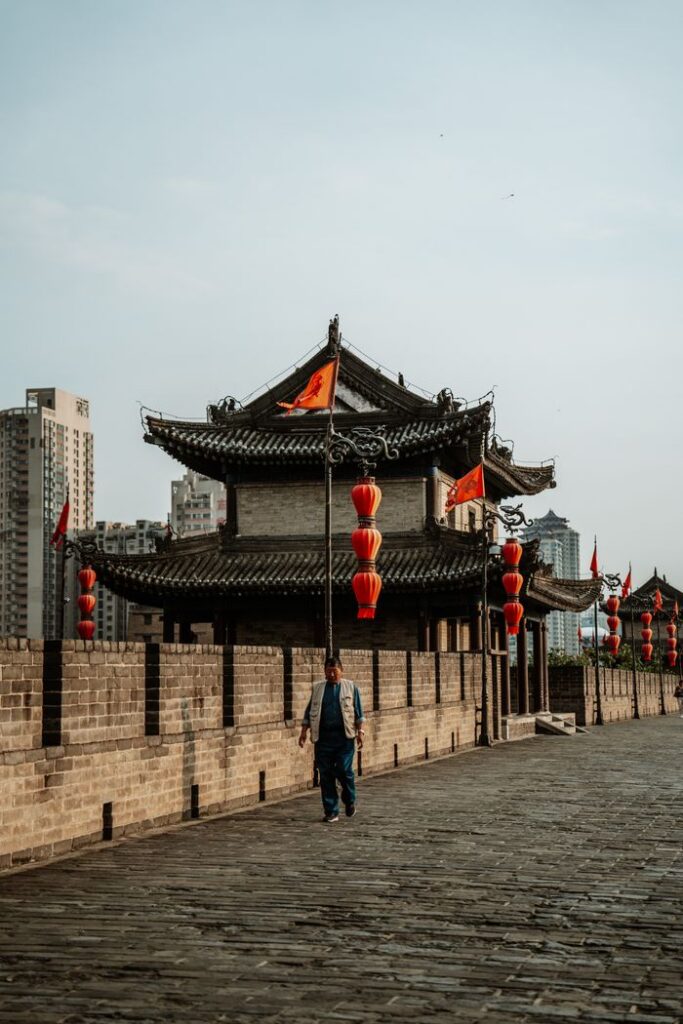 A man walks along an ancient city wall with traditional Chinese architecture and red lanterns during the day.